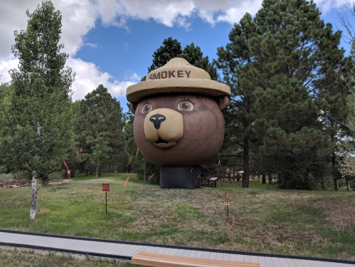 A large bear statue wearing a ranger hat stands among trees in a grassy area, with a cloudy sky above.