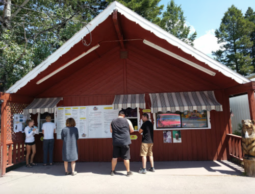 A red wooden building with a menu board, where people are ordering food under striped awnings on a sunny day.