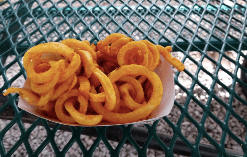 A serving of golden curly fries in a paper tray, resting on a green metal table.