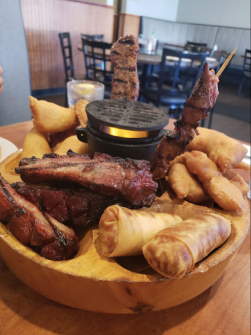 A wooden platter filled with grilled meats, spring rolls, and fried snacks, with a small grill in the center.