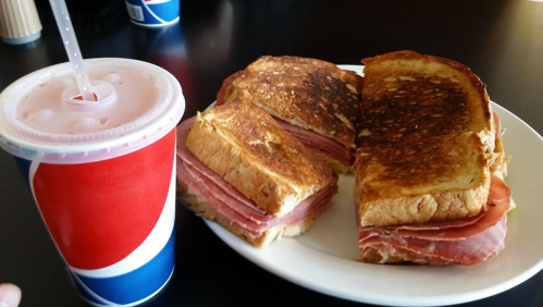 A plate with two toasted sandwiches filled with ham, alongside a cup of soda.