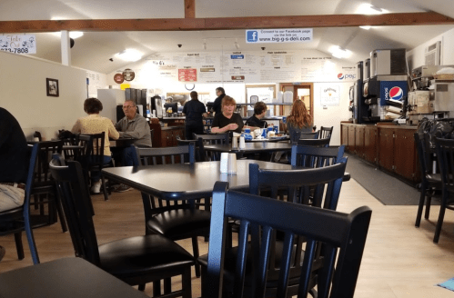 A cozy diner interior with black tables and chairs, patrons eating, and a counter in the background.