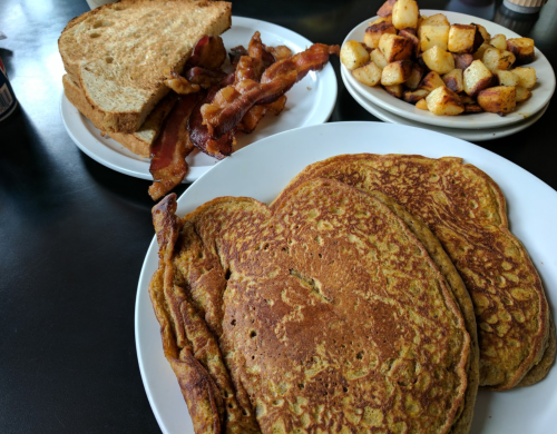 A plate of golden pancakes, crispy bacon, toast, and seasoned potato cubes on a black table.