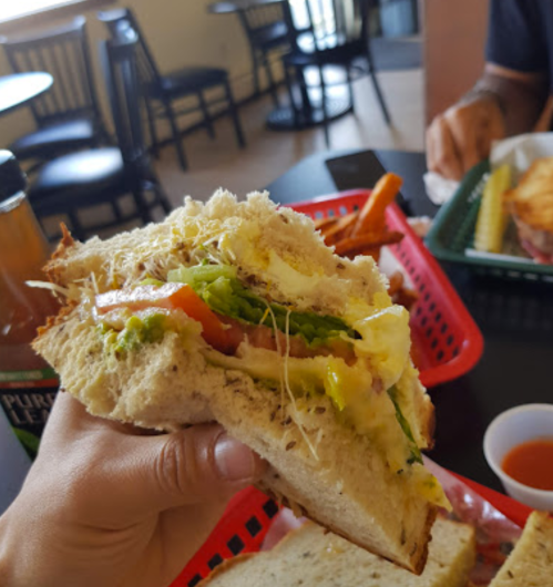A hand holding a sandwich filled with lettuce, tomato, and cheese, with fries and a drink in the background.