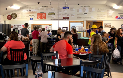 A busy restaurant scene with people ordering food at the counter and dining at tables, featuring a menu on the wall.