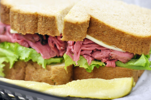 A close-up of a sandwich with layers of roast beef, lettuce, and mayonnaise, served on whole grain bread with a pickle.