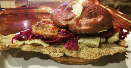 A pretzel bun filled with fried fish, red cabbage, and a tangy sauce, served on a wooden tray.