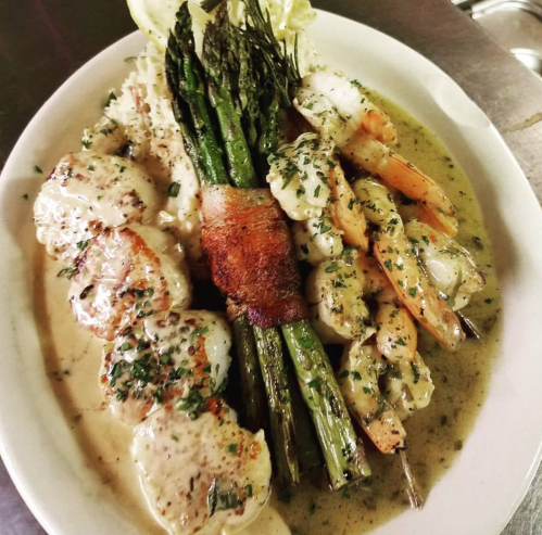 A plate of grilled shrimp and scallops, wrapped in bacon, served with asparagus and a creamy sauce.