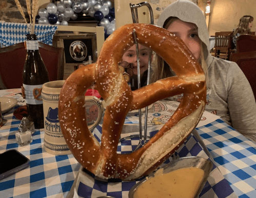 A large pretzel on a table with a beer bottle and a dipping sauce, with two children peeking from behind it.