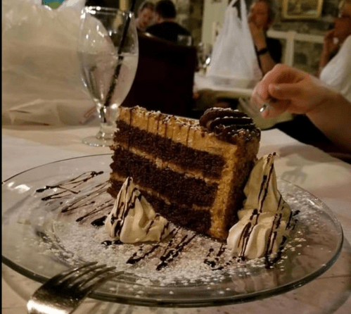 A slice of layered chocolate cake with frosting, drizzled with chocolate sauce, on a glass plate.