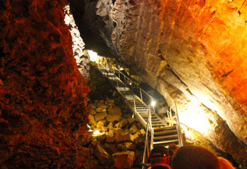 A dimly lit cave with a staircase leading upward, surrounded by rocky walls and illuminated by warm lights.