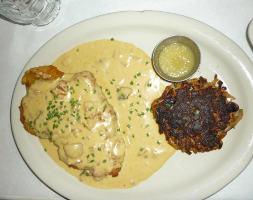 A plate featuring fried chicken smothered in creamy gravy, served with a side of crispy potato cake and applesauce.
