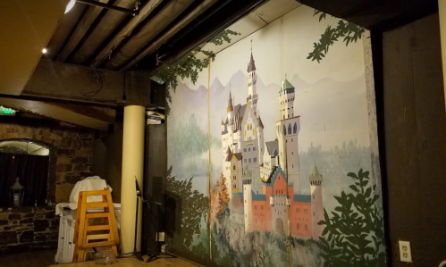 A mural of a fairytale castle surrounded by trees and mountains, set in a dimly lit room with wooden beams.