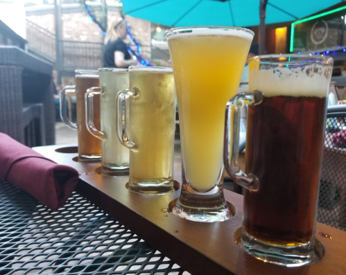 A flight of five beer samples on a table, featuring various colors and styles, with a patio setting in the background.