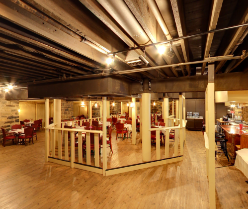 Interior of a rustic restaurant with wooden floors, tables, and a bar area, featuring exposed beams and warm lighting.