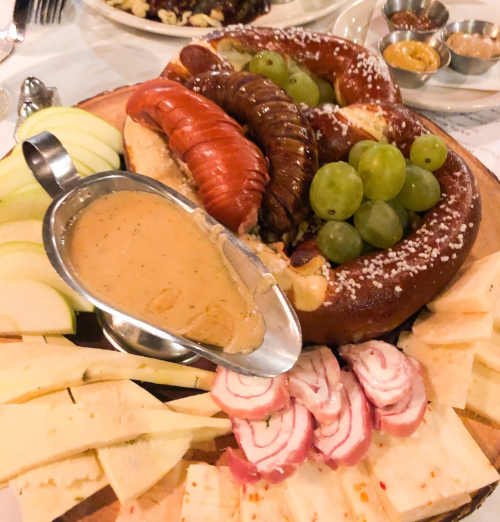 A wooden platter with a large pretzel, cheese, sliced apples, grapes, and a bowl of dipping sauce.
