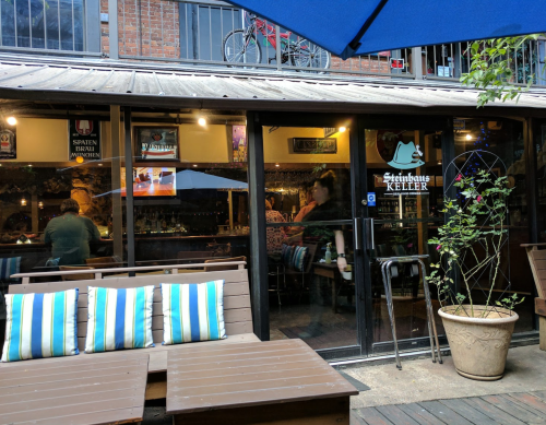 A cozy outdoor seating area in front of a bar with blue umbrellas and striped cushions, featuring a welcoming entrance.