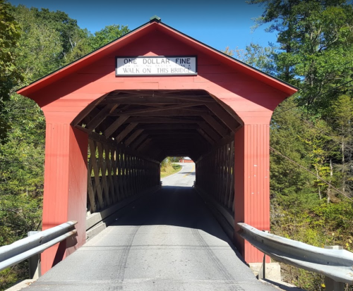 A red covered bridge with a sign stating "One Dollar Fine Walk on This Bridge," surrounded by greenery.