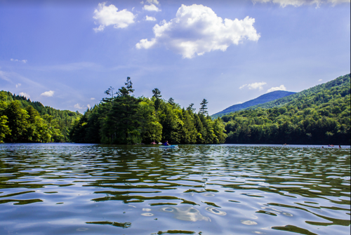 A serene lake surrounded by lush green trees and mountains under a blue sky with fluffy clouds.