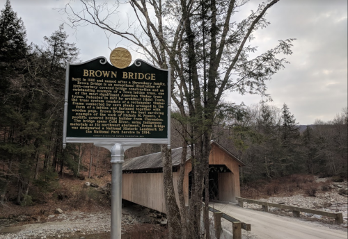 Historic sign for Brown Bridge, with a covered wooden bridge and trees in the background.