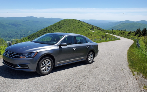 A gray sedan parked on a scenic mountain road with lush green hills and a clear blue sky in the background.
