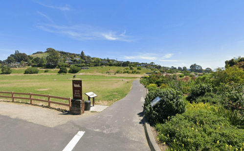 A scenic park entrance with a trail sign, grassy fields, and distant hills under a clear blue sky.