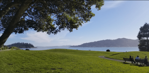 A scenic view of a grassy park by the water, with people sitting on a bench and hills in the background.