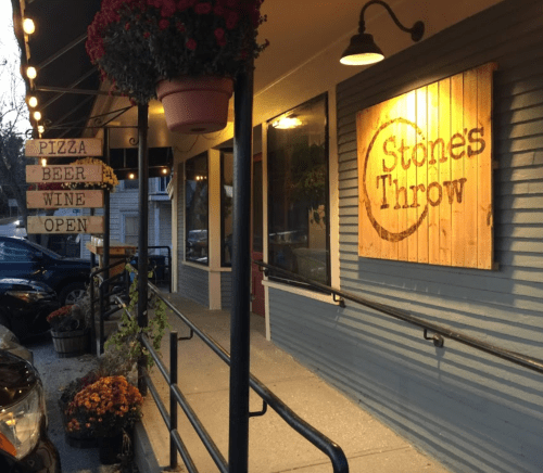 Exterior of Stones Throw restaurant with signs for pizza, beer, and wine, and flower pots lining the entrance.