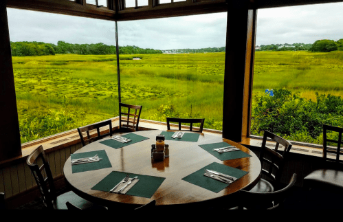 A dining table set for a meal, with large windows showcasing a lush green marshland outside.