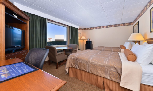 A hotel room with two beds, a desk, a TV, and a window with natural light. Decor includes curtains and artwork.