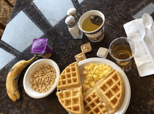 A breakfast spread with waffles, scrambled eggs, Cheerios, a banana, tea, and condiments on a table.