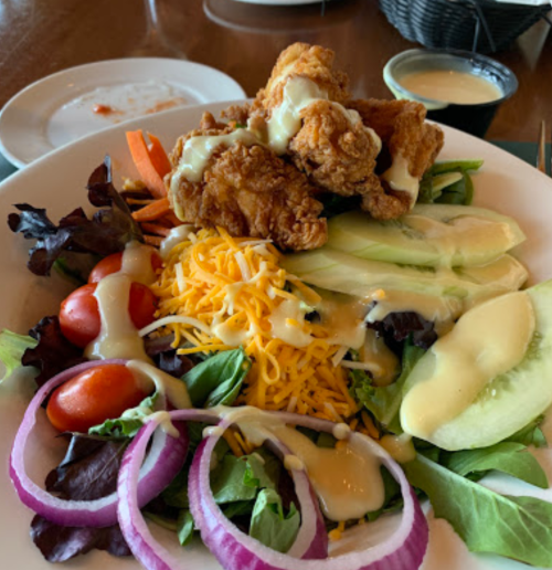 A colorful salad topped with fried chicken, shredded cheese, cucumbers, tomatoes, and drizzled with dressing.