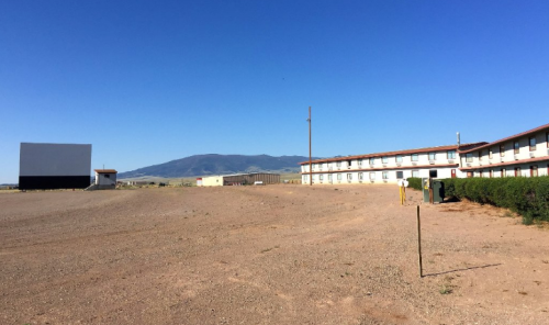 A wide view of a desert landscape featuring a drive-in movie screen, a motel, and distant mountains under a clear blue sky.