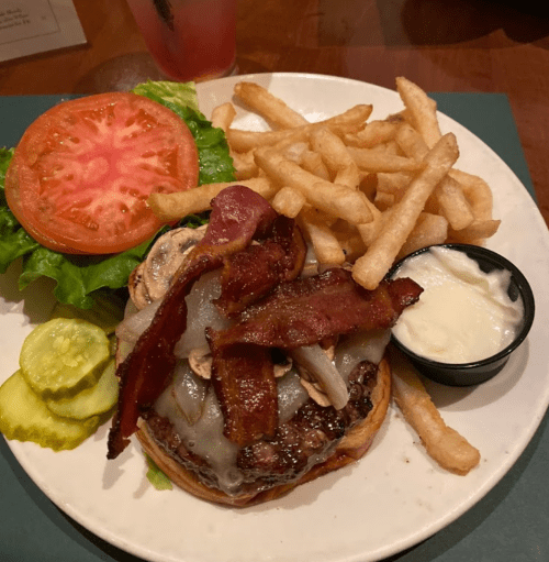 A plate with a burger topped with bacon, mushrooms, and cheese, served with lettuce, tomato, pickles, and fries.