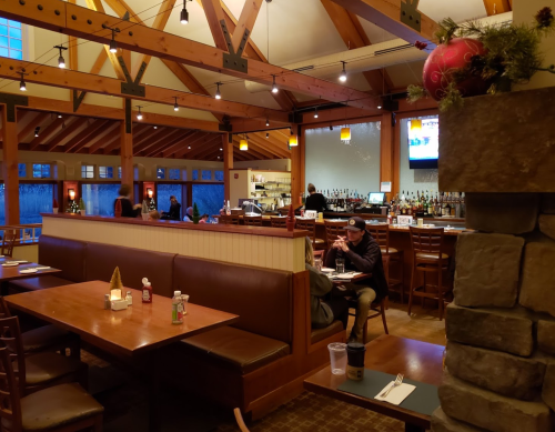 Cozy restaurant interior with wooden beams, tables, and a bar area; patrons dining and enjoying the ambiance.
