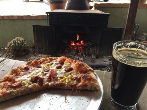 A slice of pepperoni pizza on a plate next to a glass of dark beer, with a cozy fire burning in the background.