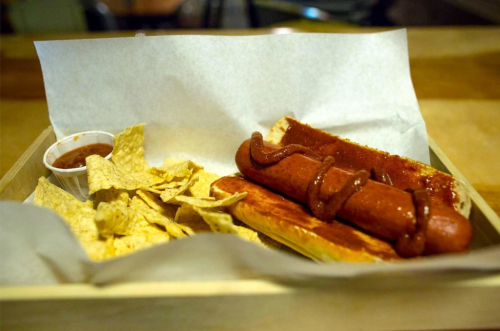 A wooden tray with two hot dogs topped with sauce, served with tortilla chips and a small cup of salsa.