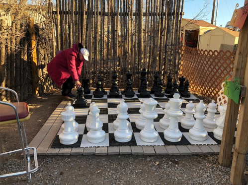 A person in a red coat adjusts a large outdoor chess piece on a giant chessboard surrounded by wooden fencing.