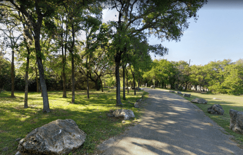 A winding path through a lush green park, lined with trees and large rocks under a clear blue sky.