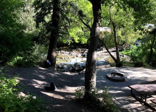 A serene outdoor scene with a rocky stream, surrounded by trees and a fire pit on a gravel area.