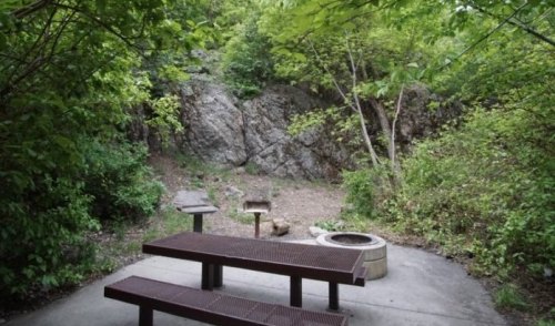 A picnic area with a table and fire pit, surrounded by lush greenery and rocky terrain.