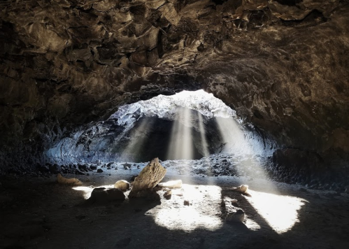 Light beams filter through a cave opening, illuminating rocky interior and scattered stones on the ground.