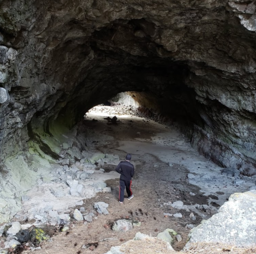 A person walks through a rocky cave, with light illuminating the entrance and scattered stones on the ground.
