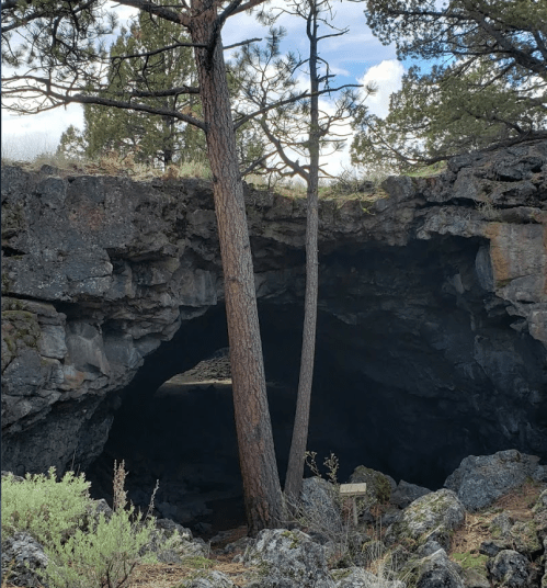 A natural cave entrance surrounded by trees and rocky terrain under a partly cloudy sky.