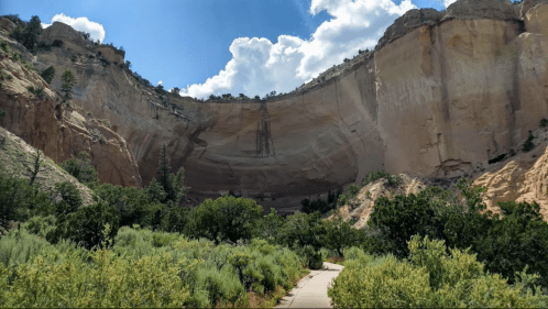 A scenic view of a large rock formation with a cave-like opening, surrounded by greenery and blue skies with clouds.