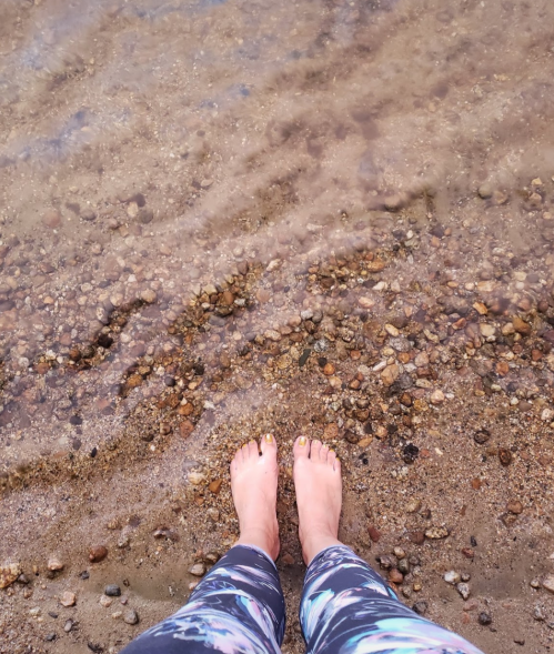 Person's bare feet standing on a rocky shore with water gently lapping around them.