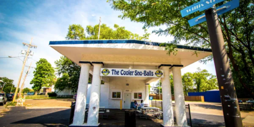 A colorful snowball stand with a blue sky, surrounded by trees and street signs, inviting customers to enjoy treats.
