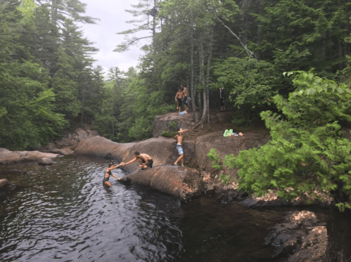 A group of people enjoying a natural swimming area, jumping off rocks into a serene, wooded setting.
