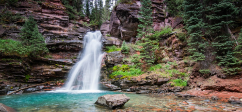 A serene waterfall cascades into a clear pool, surrounded by lush greenery and rocky cliffs.