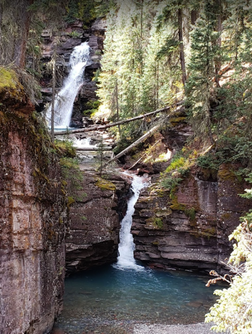 A serene landscape featuring two waterfalls cascading into a turquoise pool, surrounded by lush green trees and rocky cliffs.